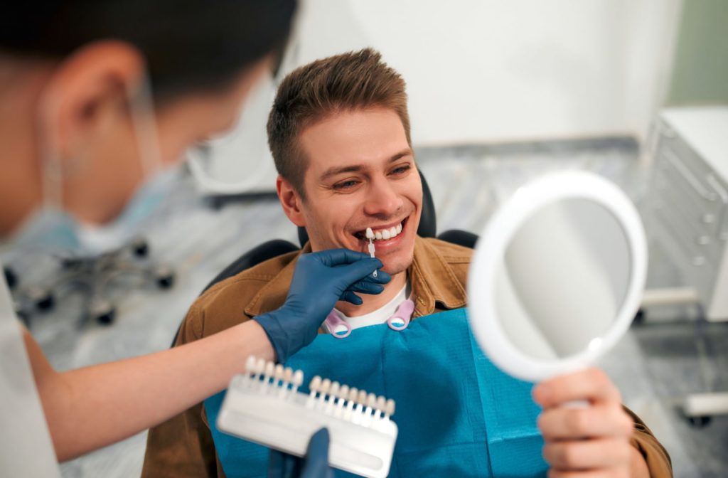 A young man looks at different shades of veneers in a small hand mirror at the dentist's office