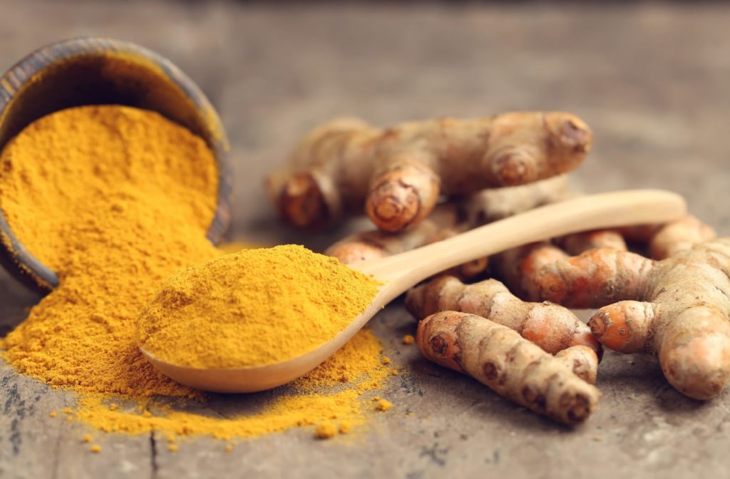 Turmeric powder spilled out onto a table, sitting next to a wooden spoon that has turmeric powder in it. The powder is also surrounded by several turmeric plant stems