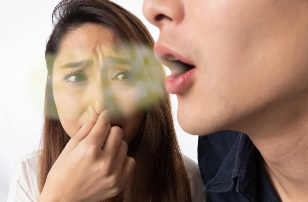 A person pinches her nose shut while another person is speaking, to protect herself from the speaker's bad breath. A stylised green and white cloud comes out of the speaker's mouth to highlight the smell of their breath.