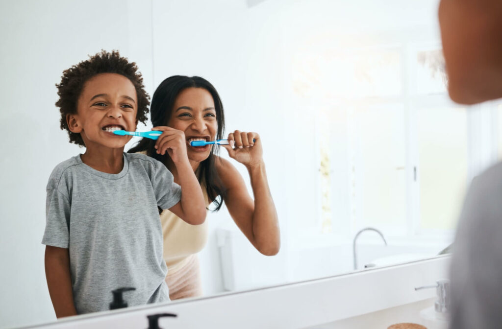 A child and their mother stand in front of a mirror in a brightly-lit bathroom, brushing their teeth together.