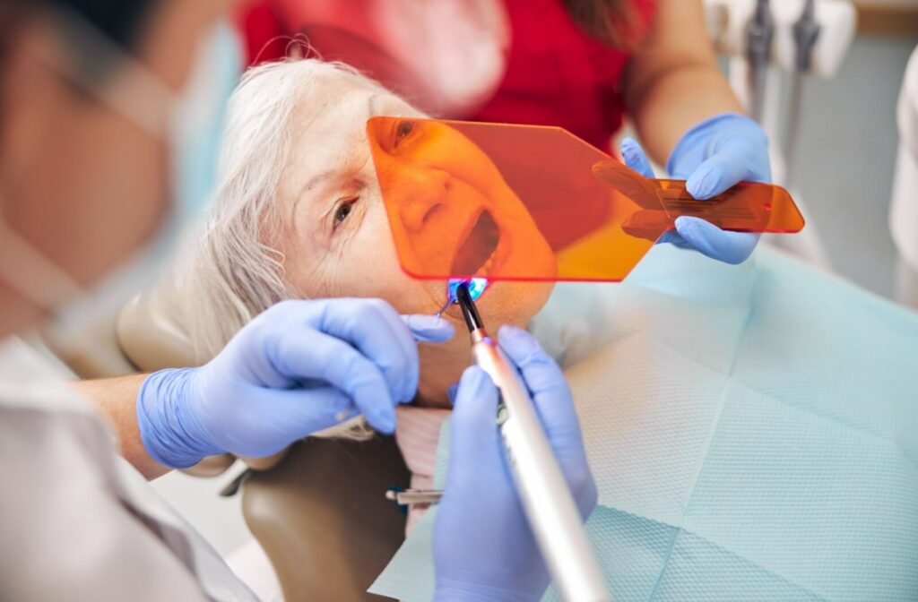 A patient lies in a dentist's chair while the dentist performs a dental bonding procedure. A dental hygienist holds a plastic screen over the patient's face to expose the bonding resin to UV light.