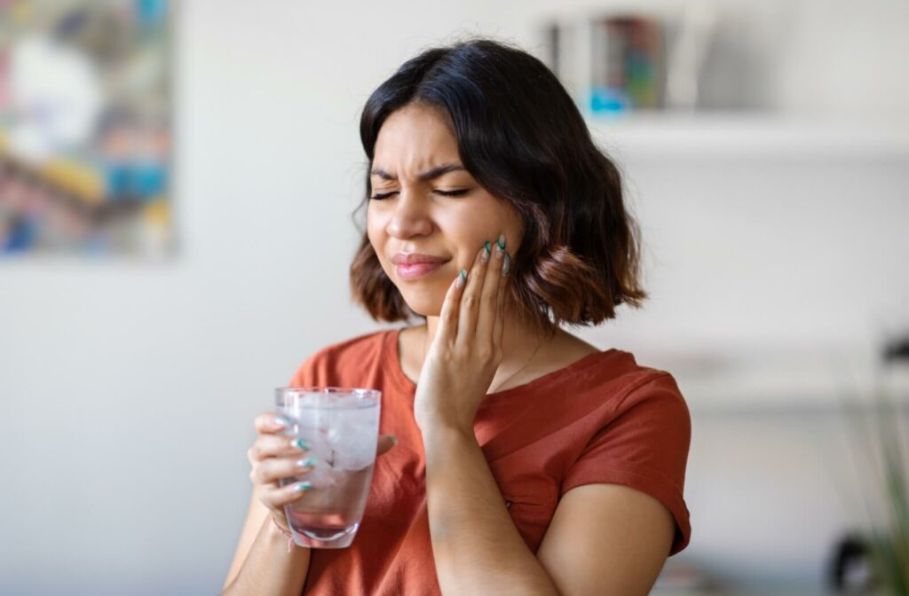 A young woman rubbing her cheek in discomfort while holding a glass of ice water after a tooth extraction.