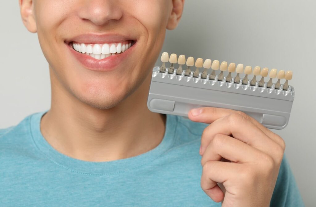 A person smiles widely with very white teeth while holding a veneer tooth shade tablet next to their mouth.