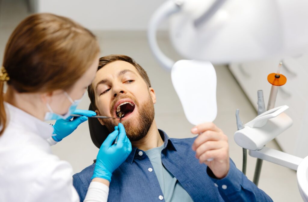 A dental patient in a blue shirt holds up a mirror to watch as a dental hygienist cleans plaque off their teeth