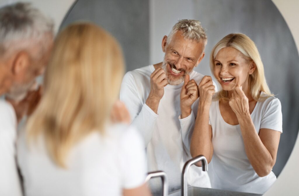 A smiling senior couple in matching white shirts looks into their bathroom mirror as they floss their teeth