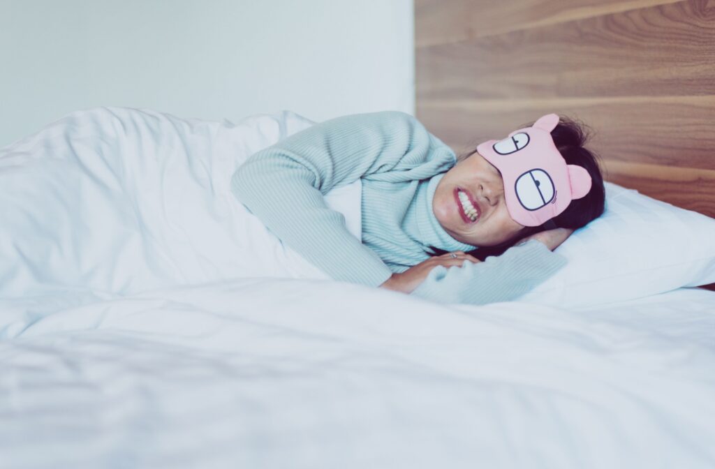 An adult lays in a clean, cozy bed with a sleep mask on, clenching and grinding their teeth.