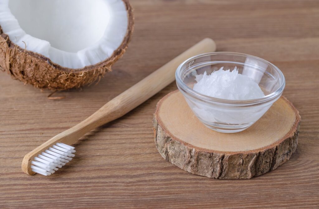 Toothbrush and bowl of coconut oil on wooden surface next to half a coconut.