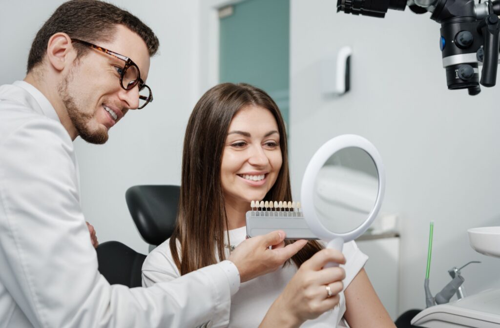 Smiling woman holding a mirror while dentist shows veneer shade options during a cosmetic consultation.