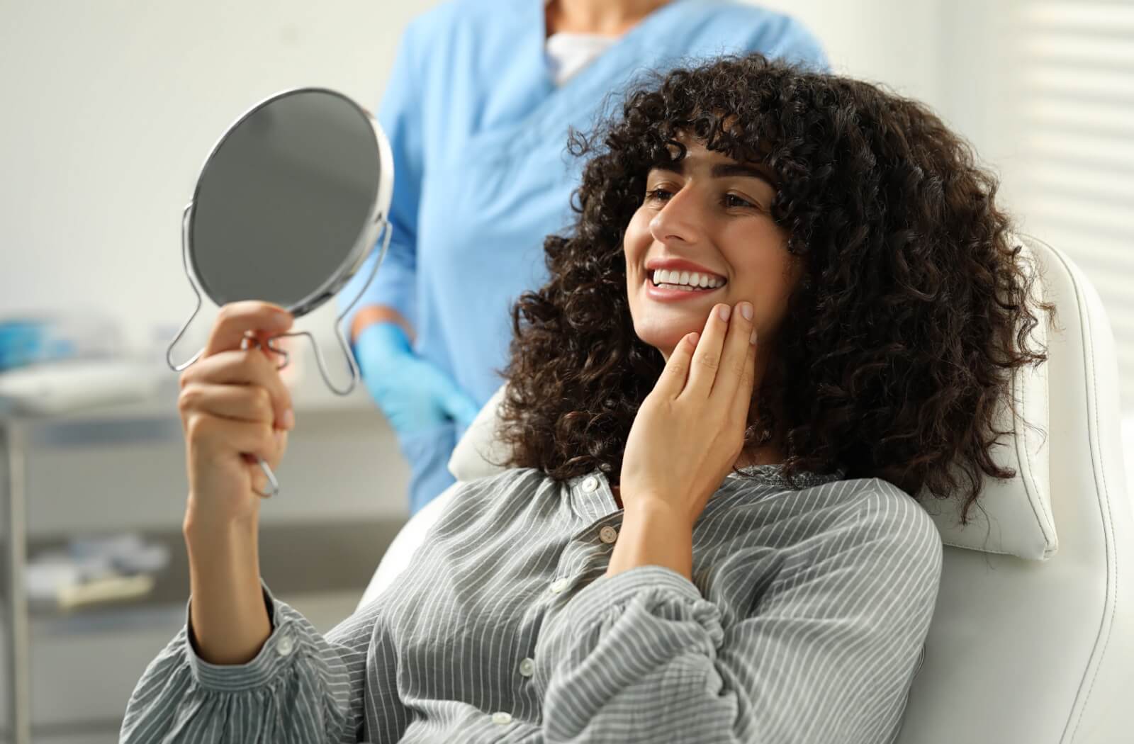 Woman smiling in dental chair while examining her veneers with a handheld mirror after a cosmetic dental procedure.