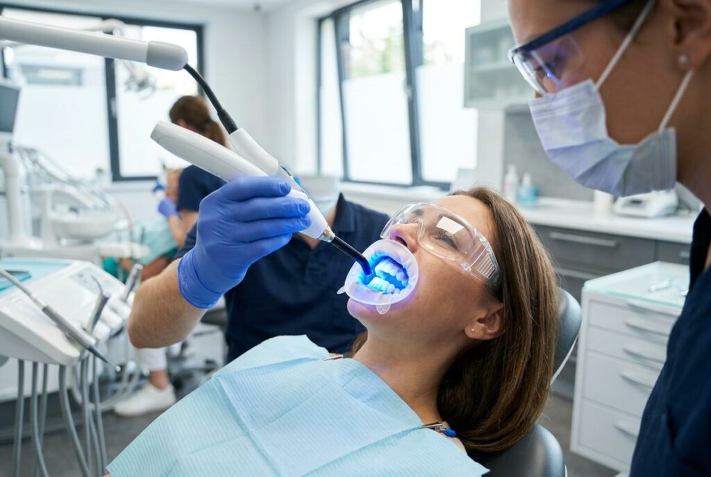 Dentist applies a blue UV light tool to a seated patient's open mouth in a dental clinic. Patient wears protective goggles.